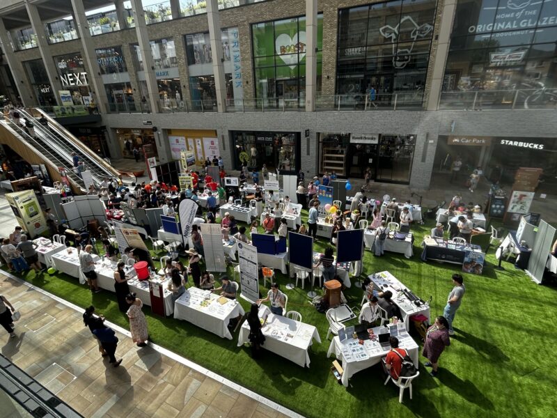 A group of people surrounding exhibition stands in a shopping centre.