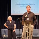 A man dressed in a black shirt talks into a mic. A lady stands behind him.