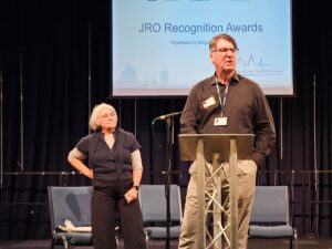 A man dressed in a black shirt talks into a mic. A lady stands behind him.