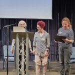 Three people stand on stage looking at a printed, framed certificate.